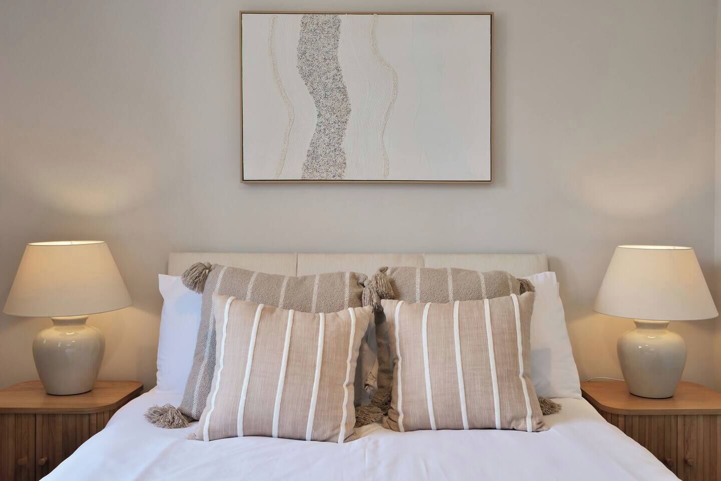 Una cama bien hecha con almohadillas a rayas y dos lámparas de noche sobre mesitas de madera.