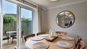 A dining area with a wooden table, woven placemats, and a candle centerpiece. A round mirror hangs on the wall, and a sliding glass door leads to an outdoor patio with a view of a garden and a wooden fence.