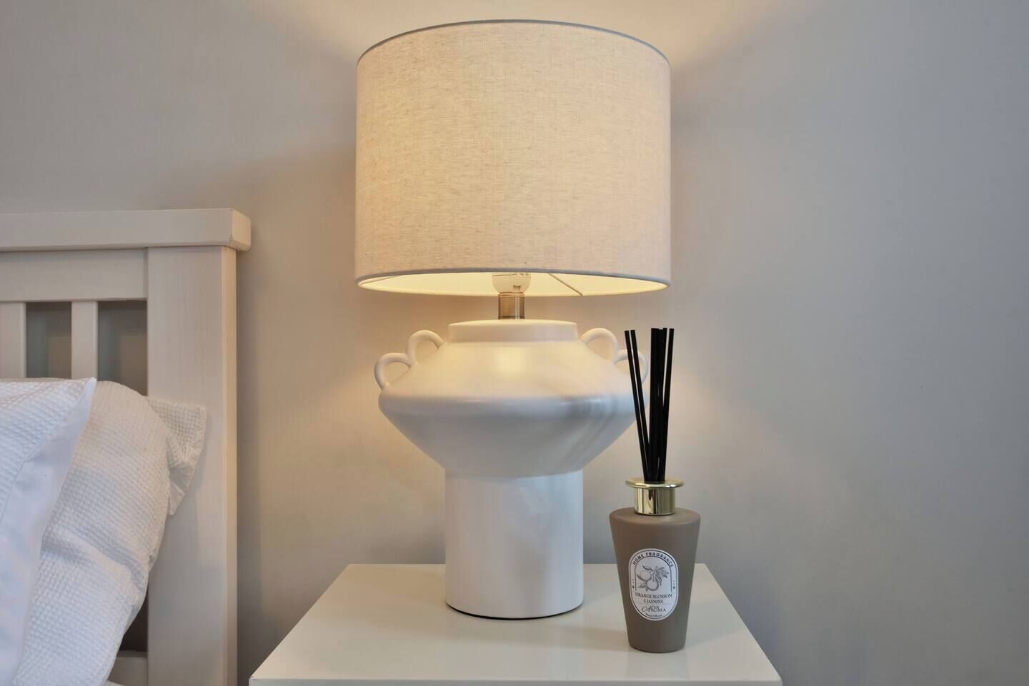 Een nachtkastje met een witte lamp, een geribbelde lampenkap en een decoratieve vaas met zwarte takken.