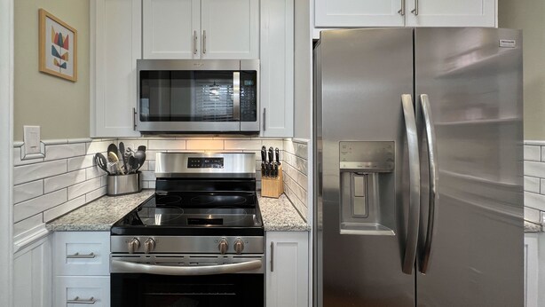 A kitchen with white cabinets, a stainless steel refrigerator, a microwave, and a stove. - Modern Hickory Hideaway (Hickory)