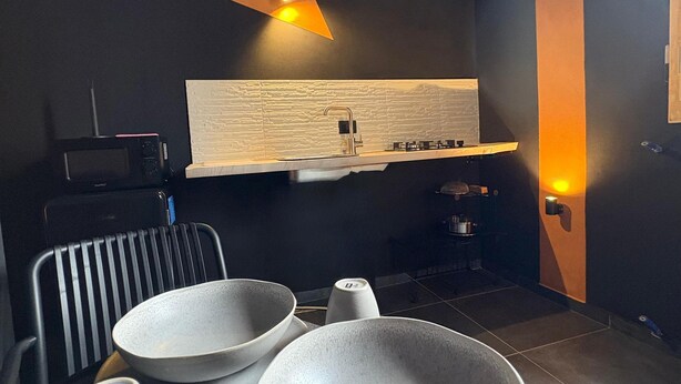 Ett modernt kök med en mörk vägg, ett träpanelat tak och en lampa ovanför bänkskivan. På bänkskivan står två skålar, en vattenkokare och ett litet bord med en stol.