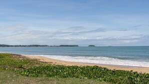 A beach with a sandy shore and green vegetation in the foreground. - Apto com Wi-fi a 950m da Praia da Tartaruga (Rio das Ostras)