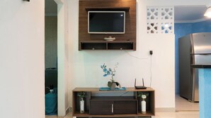 A modern living room with a flat-screen TV mounted on a wooden wall, a dark wooden console table with decorative items, and a built-in refrigerator. - Apto com Wi-fi a 950m da Praia da Tartaruga (Rio das Ostras)