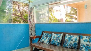 A room with a blue wall, a wooden bench with floral cushions, and a view of greenery through a window. - Apto com Wi-fi a 950m da Praia da Tartaruga (Rio das Ostras)
