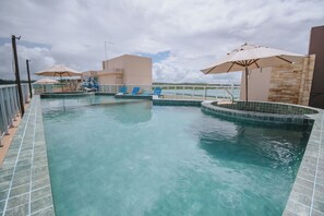 A swimming pool with clear blue water, surrounded by lounge chairs and umbrellas, and a view of the sky and distant landscape. - Flat com Wifi a 400m da Praia de Porto de Galinhas (Ipojuca)