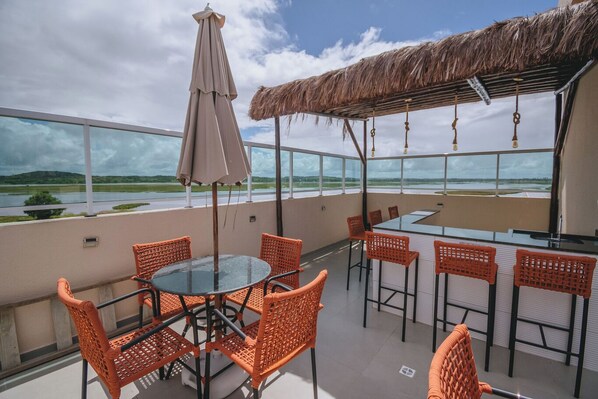 Una terraza en la azotea con una mesa de vidrio, sillas de mimbre color naranja, un techo de paja y vistas a una masa de agua.