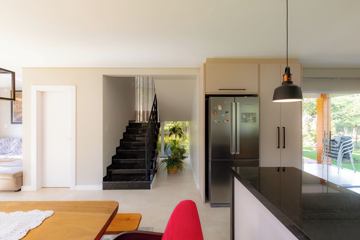 Una cocina moderna con un refrigerador de acero inoxidable, un área de comedor con una mesa de madera y sillas rojas, y una escalera que lleva a un balcón con plantas.