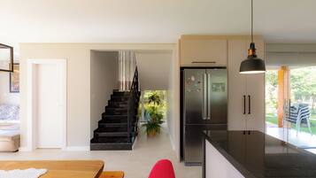 Una cocina moderna con un refrigerador de acero inoxidable, un área de comedor con una mesa de madera y sillas rojas, y una escalera que lleva a un balcón con plantas.