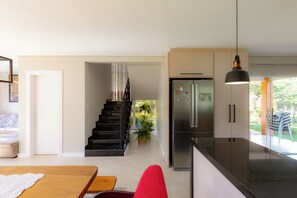A modern kitchen with a stainless steel refrigerator, a dining area with a wooden table and red chairs, and a staircase leading to a balcony with plants.