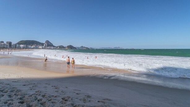 Ein Strand mit Menschen, die am Ufer entlanggehen, feiner Sand und ein klarer, blauer Himmel.