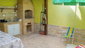 Cucina con forno a mattoni, lavello e piccolo bancone con barattoli. Si nota una sedia da spiaggia colorata e un ombrello verde.