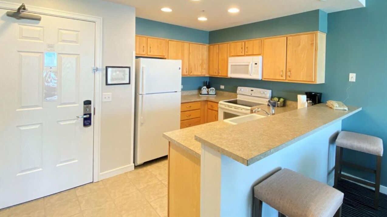 A modern kitchen with wooden cabinets, a white refrigerator, a microwave, and a stove. There are two bar stools and a small island with a sink.