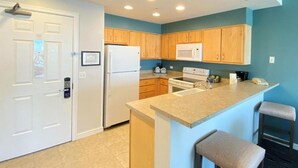 A modern kitchen with wooden cabinets, a white refrigerator, a microwave, and a stove. There are two bar stools and a small island with a sink. - Two Bedroom KIng WorldMark Seaside (Seaside)
