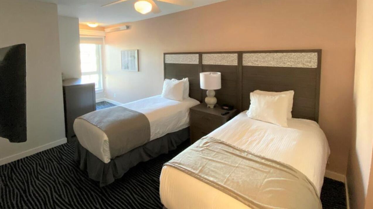 Quarto de hotel com duas camas, uma mesinha de cabeceira com abajur, um espelho grande e uma janela com persianas.
