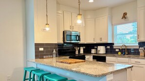 A modern kitchen with white cabinets, a granite countertop, a central island with bar stools, a stainless steel microwave, and a window with blinds.