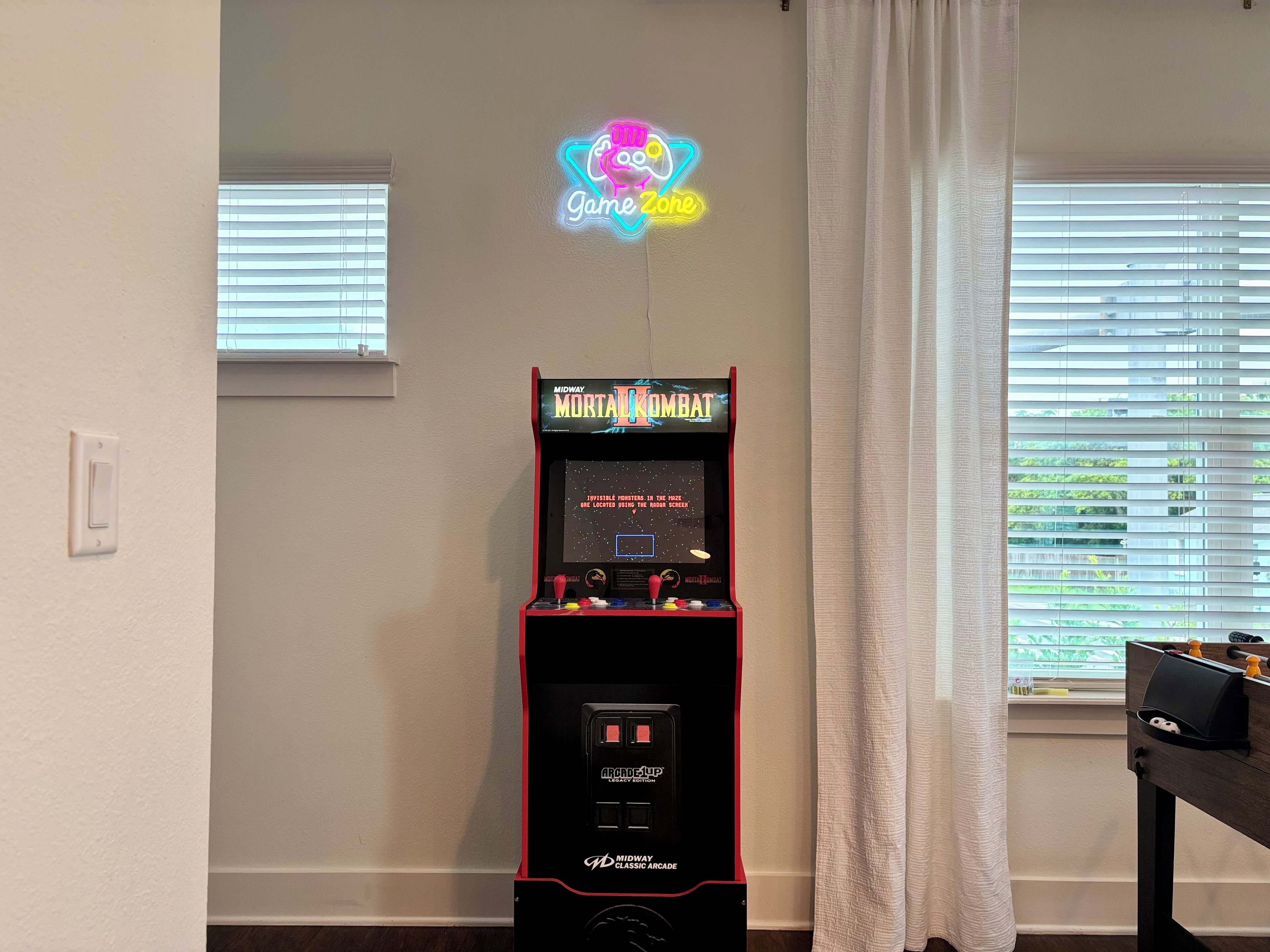 A Midway arcade machine labeled 'Mortal Kombat' in a room with a 'Game Zone' neon sign, a window with blinds, and a curtain.