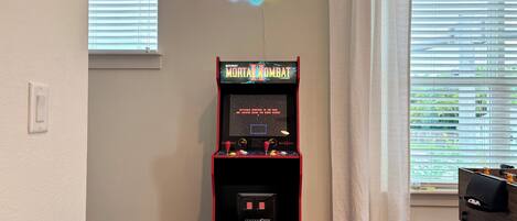 A Midway arcade machine labeled 'Mortal Kombat' in a room with a 'Game Zone' neon sign, a window with blinds, and a curtain.