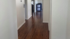 Un couloir aux murs blancs et au plancher en bois foncé, qui mène à une pièce où se trouve un lit superposé.