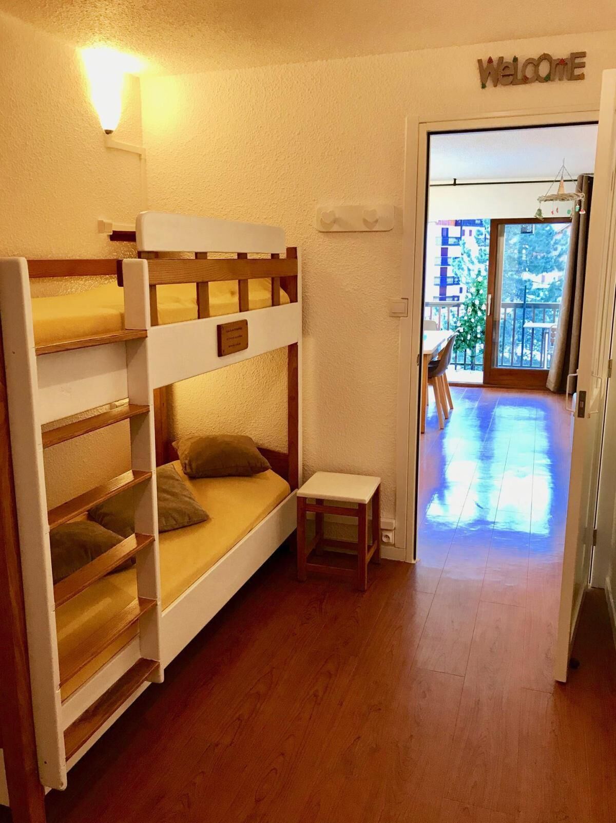A bunk bed room with a small table and a bench, a welcome sign, and a view of the outside balcony.