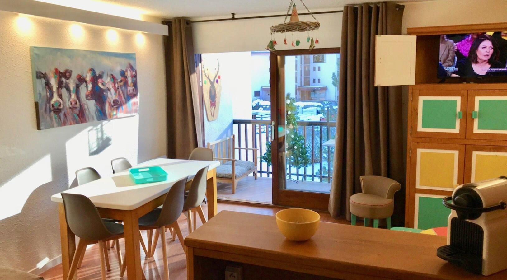 A hotel room with a dining area, a TV, a balcony with a view of the outside, and a painting on the wall.
