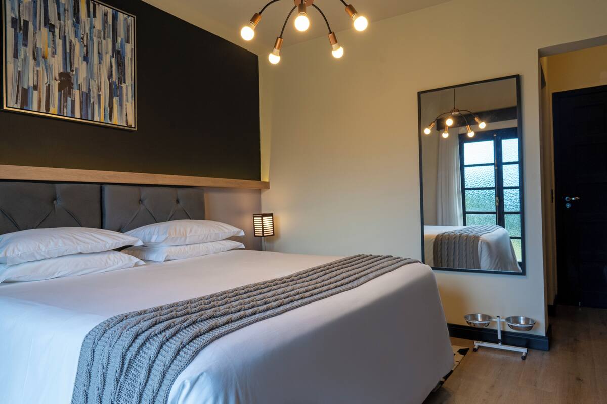 A modern bedroom with a large bed, a headboard, a bedside table, a mirror, and a window with a view.