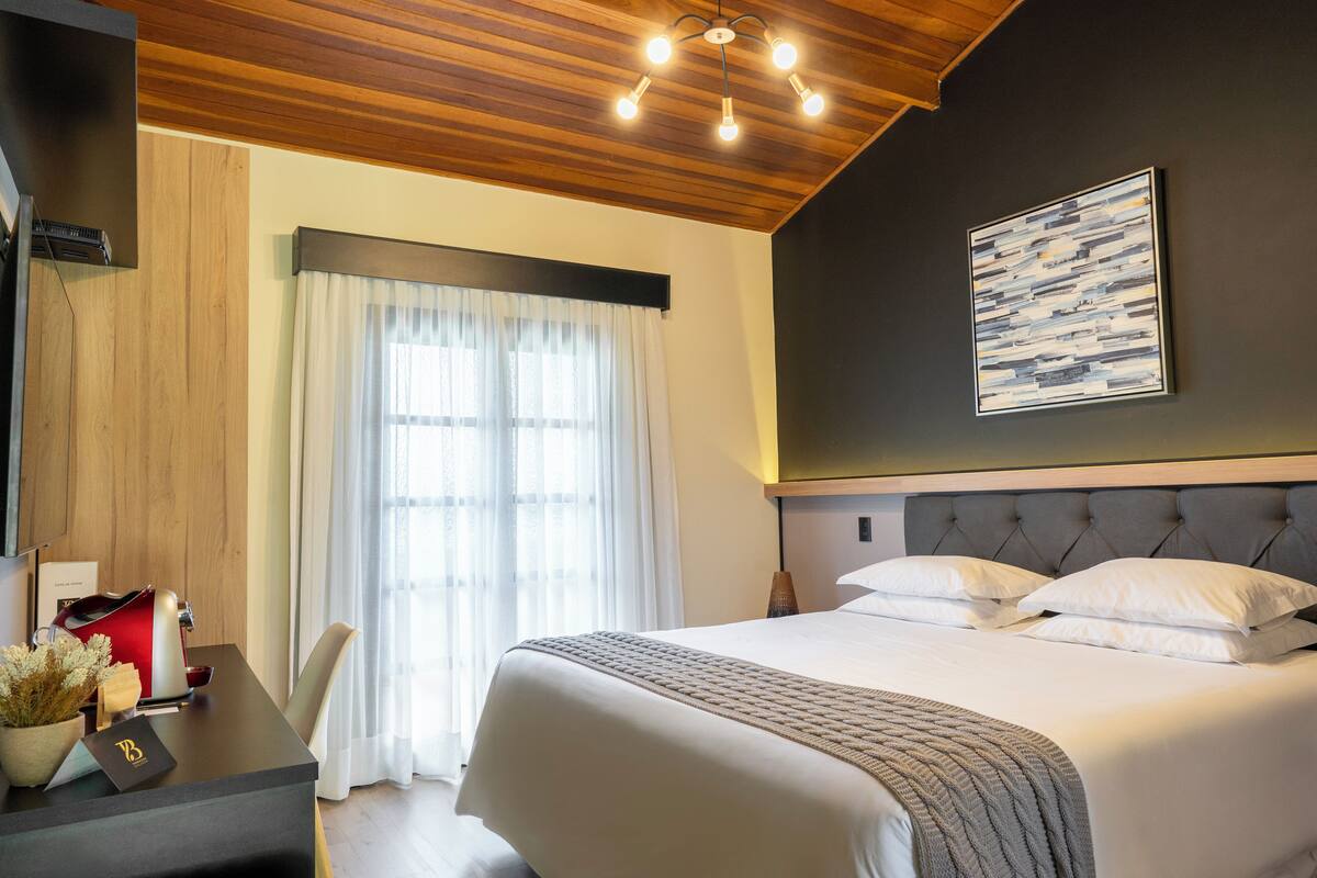 A hotel room with a large bed, a desk with a red kettle, a vase with flowers, and a painting on the wall.