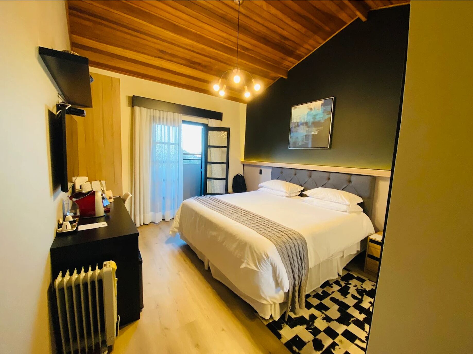A hotel room with a bed, a desk, a radiator, a door, and a window with curtains.