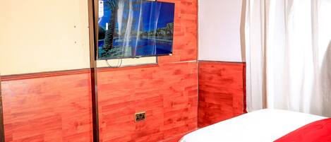 A hotel room with two beds, a large flat-screen TV mounted on the wall, and a window with white curtains.