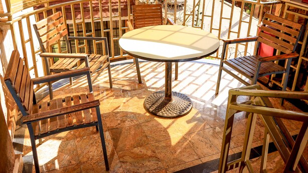 A sunlit outdoor seating area with a round table and wooden chairs, surrounded by a metal railing.