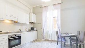 A modern kitchen with white cabinets, a built-in oven, and a microwave. There is a dining area with a table and chairs, and a window with sheer curtains.