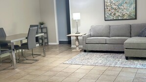 A living room with a grey sofa, a dining table with chairs, a lamp, and a painting on the wall. - 3BR Midtown Stay! Located on USM Campus!!  Perfect for Game Days and Getaways (Hattiesburg)