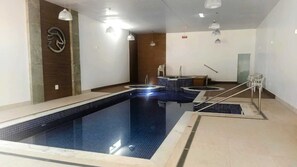 A swimming pool with a tiled edge, a wooden wall with a logo, and a ceiling with hanging lights. - Cavalinho Branco Flat Para 4 Pessoas c Piscina (Águas de Lindóia)