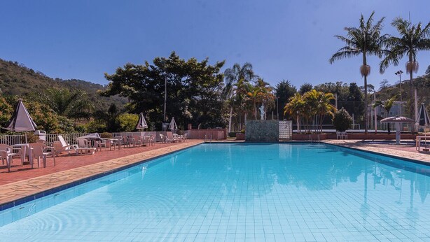 A rectangular swimming pool with tiled edges, surrounded by lounge chairs and umbrellas, set against a backdrop of trees and hills. - Cavalinho Branco Flat Para 4 Pessoas c Piscina (Águas de Lindóia)