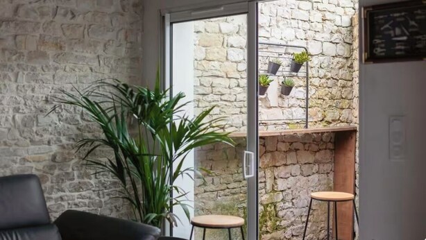 A room with a stone wall, a large plant, a sliding glass door leading to a balcony with a stone wall and potted plants. - Le Chai des Tortues · Character home · Beach 250 m (Rivedoux-Plage)