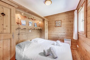 A wooden room with a bed, a shelf with books, and a window with curtains.