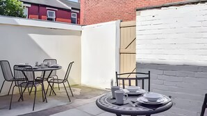 Eine Terrasse im Freien mit einem runden Tisch und Stühlen, einem weißen Zaun und einer Ziegelwand.