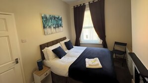 Ein Schlafzimmer mit Bett, Nachttisch, Stuhl, Schreibtisch, einem Fenster mit Vorhängen und einem Gemälde an der Wand.