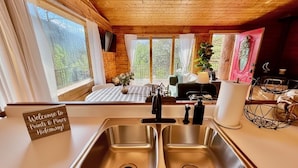 Uma cozinha com pia de aço inoxidável, um letreiro com a inscrição "Welcome to Points & Pines Hideaway!" e vista para o exterior através de grandes janelas.