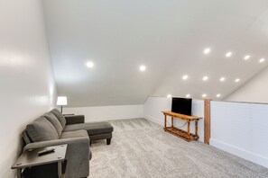 Un dormitorio estilo loft moderno con techo inclinado, un sofá seccional gris, una pequeña mesa de madera y un televisor sobre una pared blanca.
