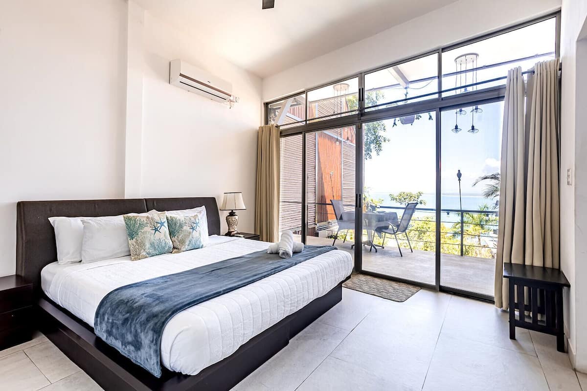 A modern hotel room with a large bed, a balcony with a view, and a sliding glass door.