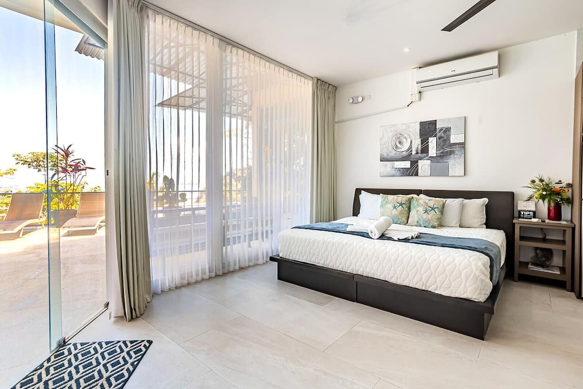 A modern bedroom with a large bed, a glass door leading to a balcony, and a wall-mounted air conditioner.