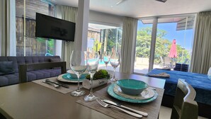 Quarto de hotel moderno com área de jantar, sofá, TV e vista para o exterior.
