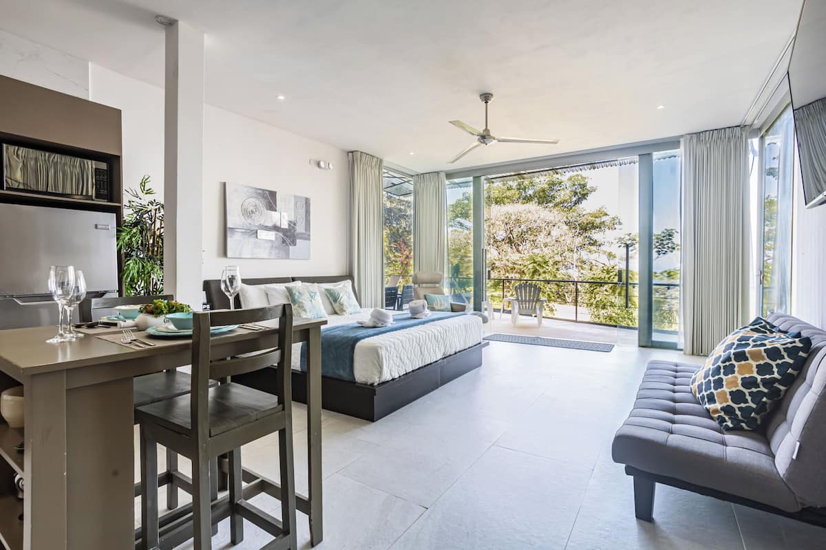 A modern bedroom with a bed, a dining area with a table and chairs, and a balcony with a view of trees.