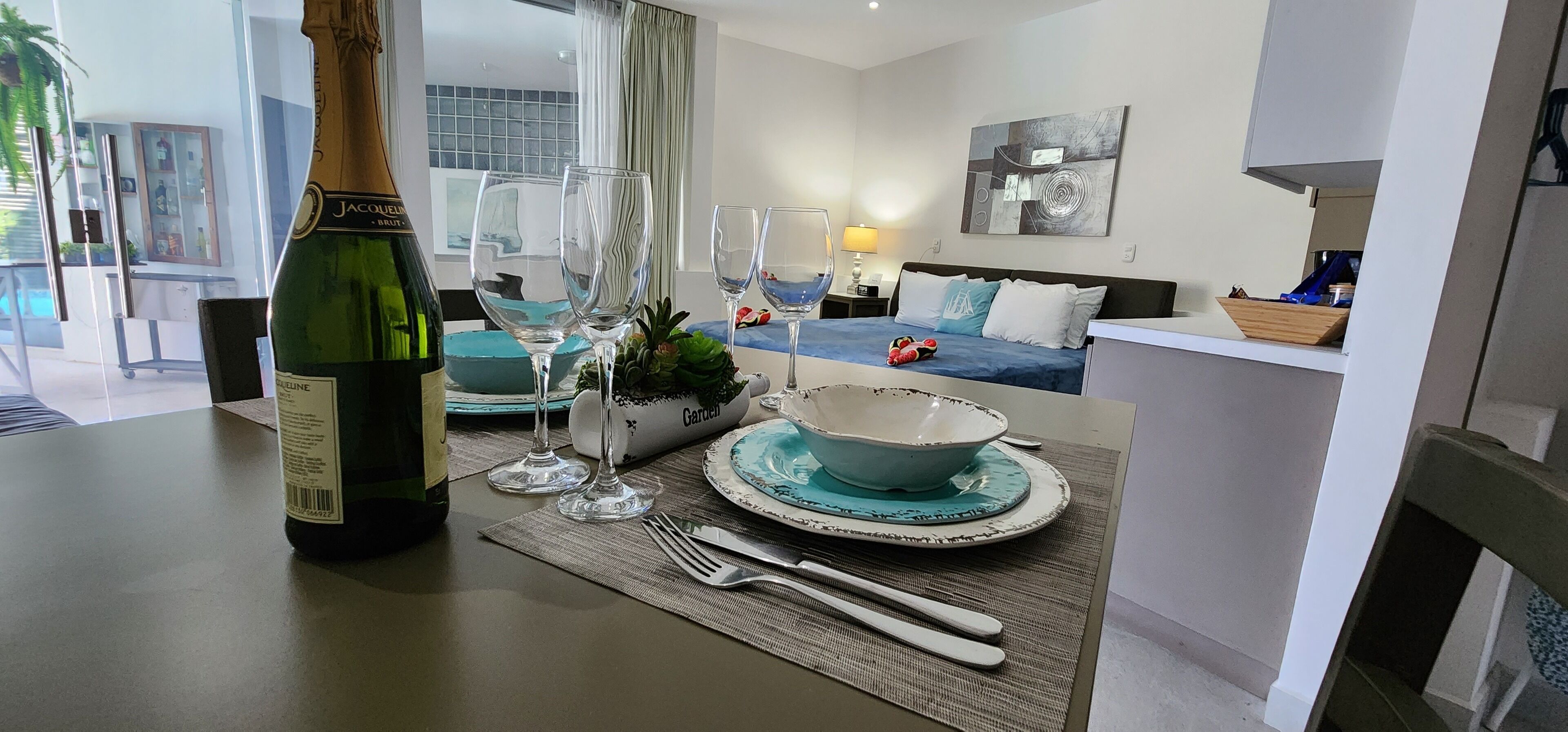 A dining table set with a bottle of champagne, glasses, and plates, in a modern living room with a sofa and artwork.