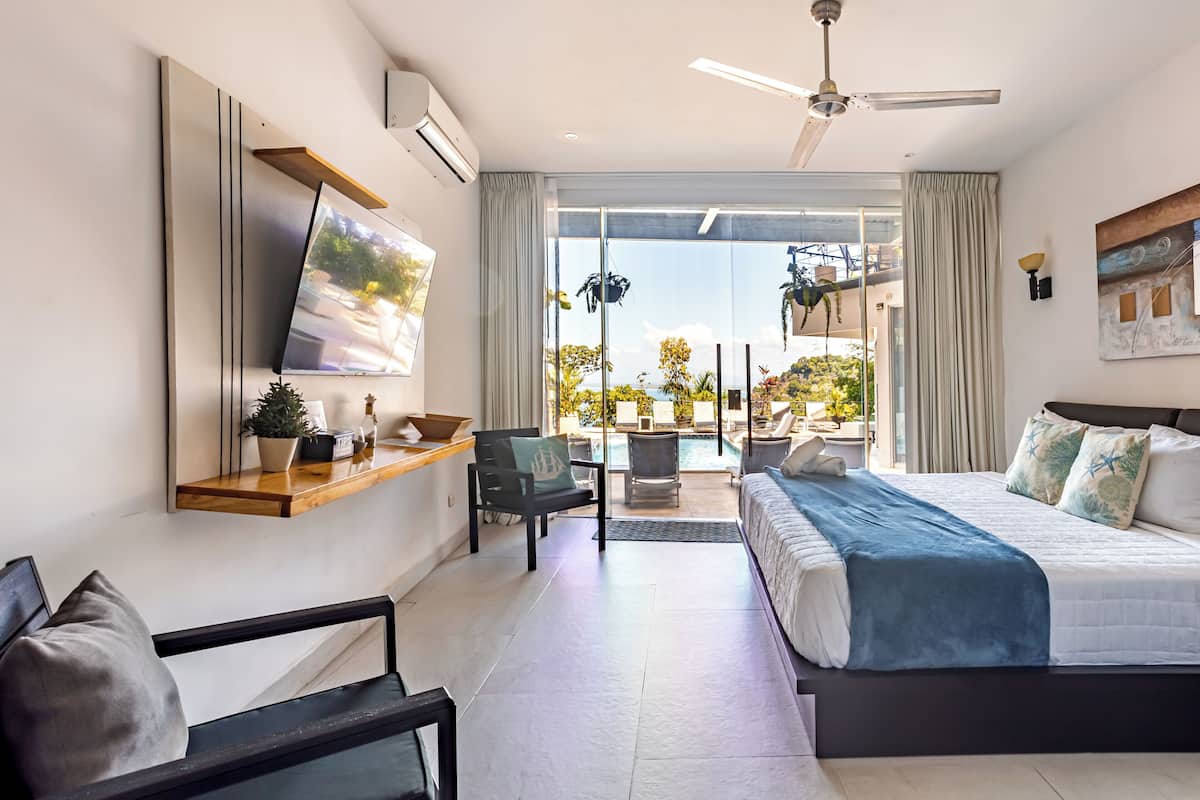 A modern hotel room with a bed, a TV, a seating area, and a view of the outdoors.