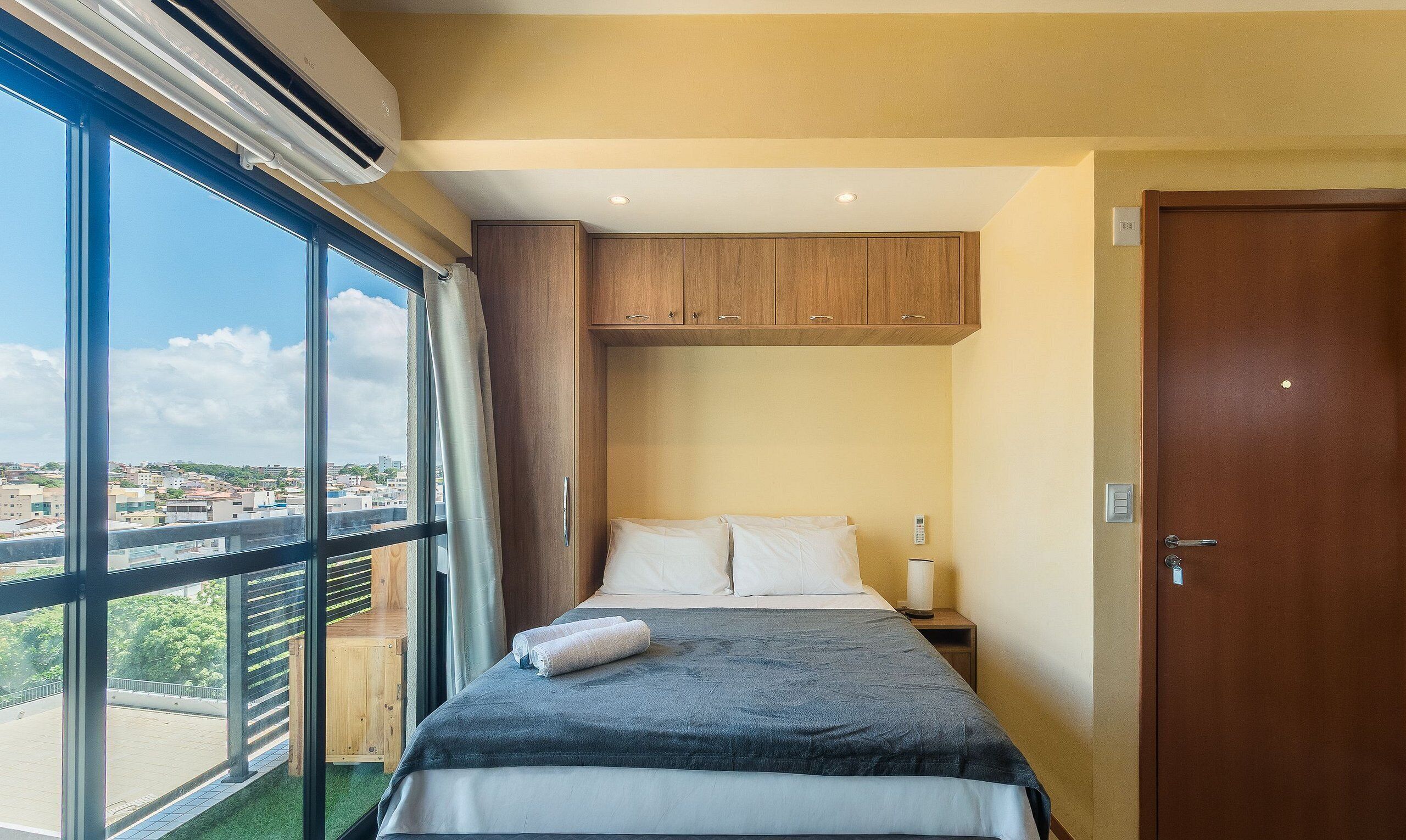 A hotel room with a large window, a bed, a wooden wardrobe, and a door.