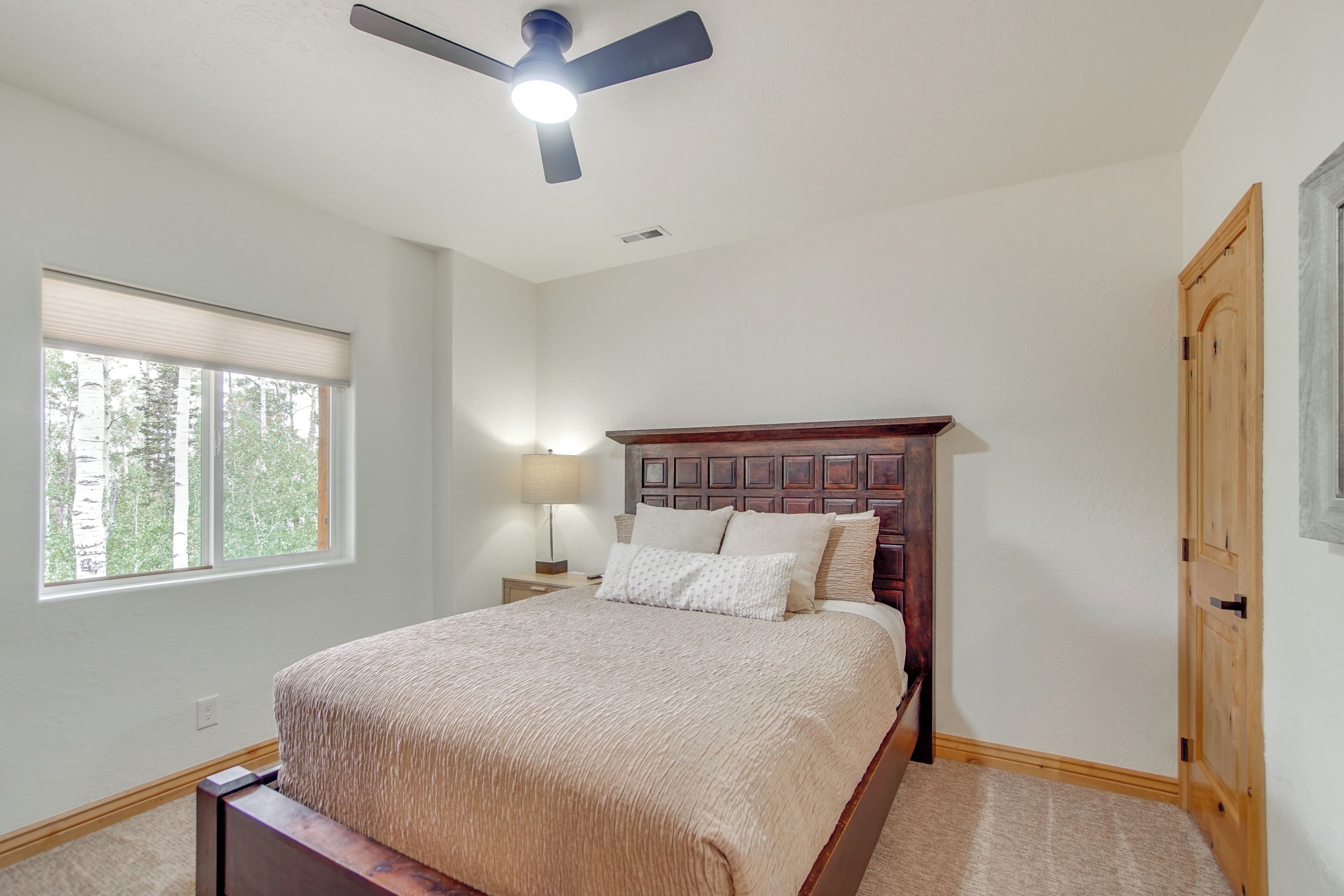 A bedroom with a bed, a ceiling fan, a nightstand with a lamp, a window, and a door.