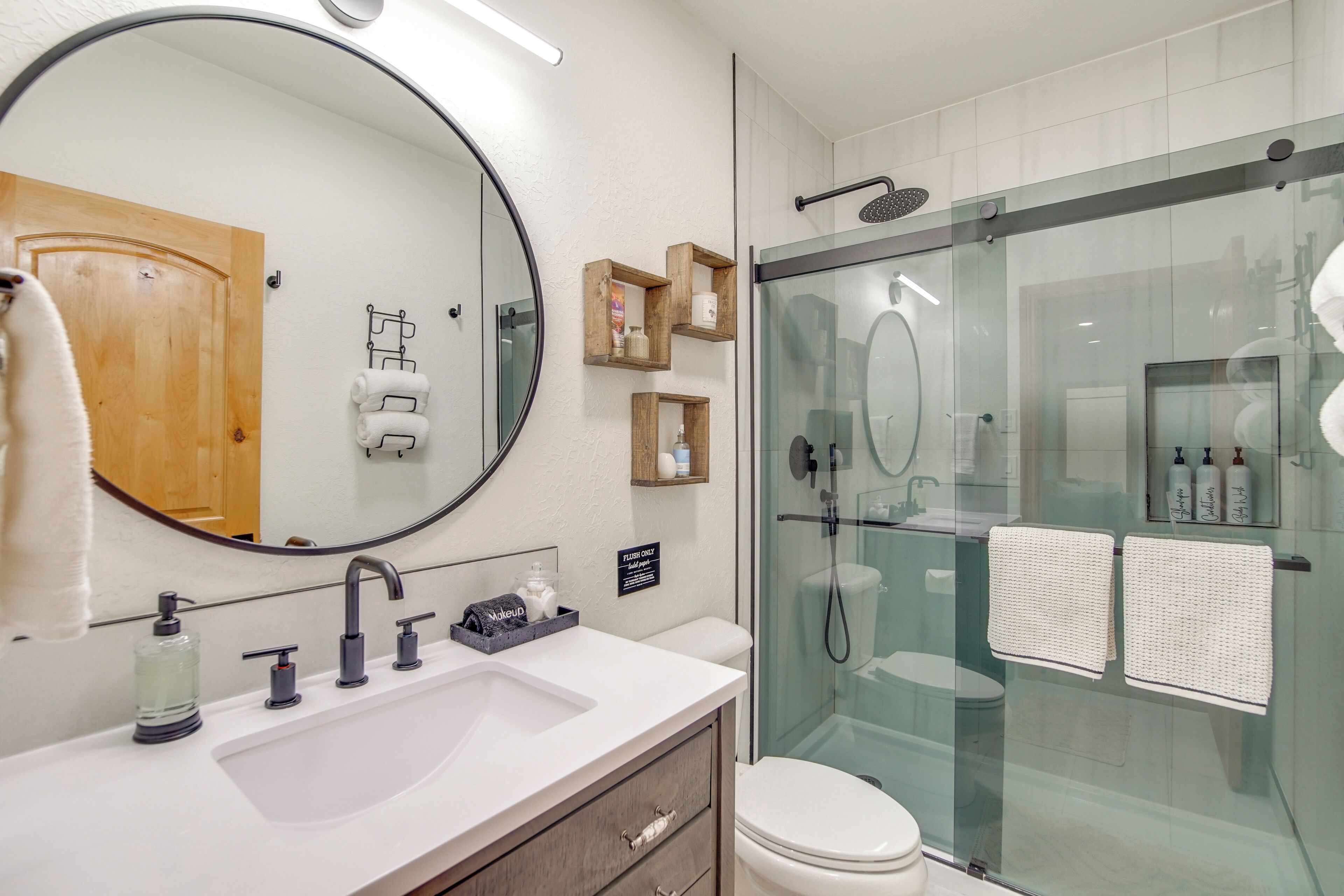 Un baño moderno con un espejo grande, una ducha con cerramiento de vidrio, un lavamanos blanco y una puerta de madera.