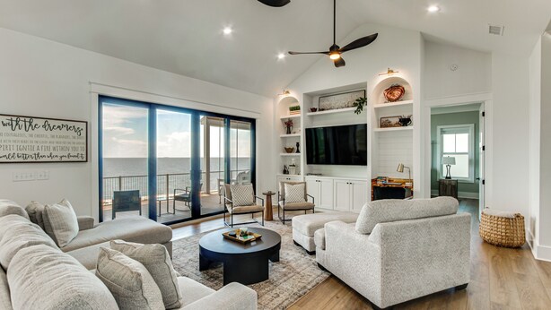 A spacious living room with a large sofa, two armchairs, a coffee table, and a flat-screen TV. There are ceiling fans, a bookshelf, and a view of the outdoors through large windows. - Luxury Beachfront Living! Epic Ocean Springs Home (Ocean Springs)