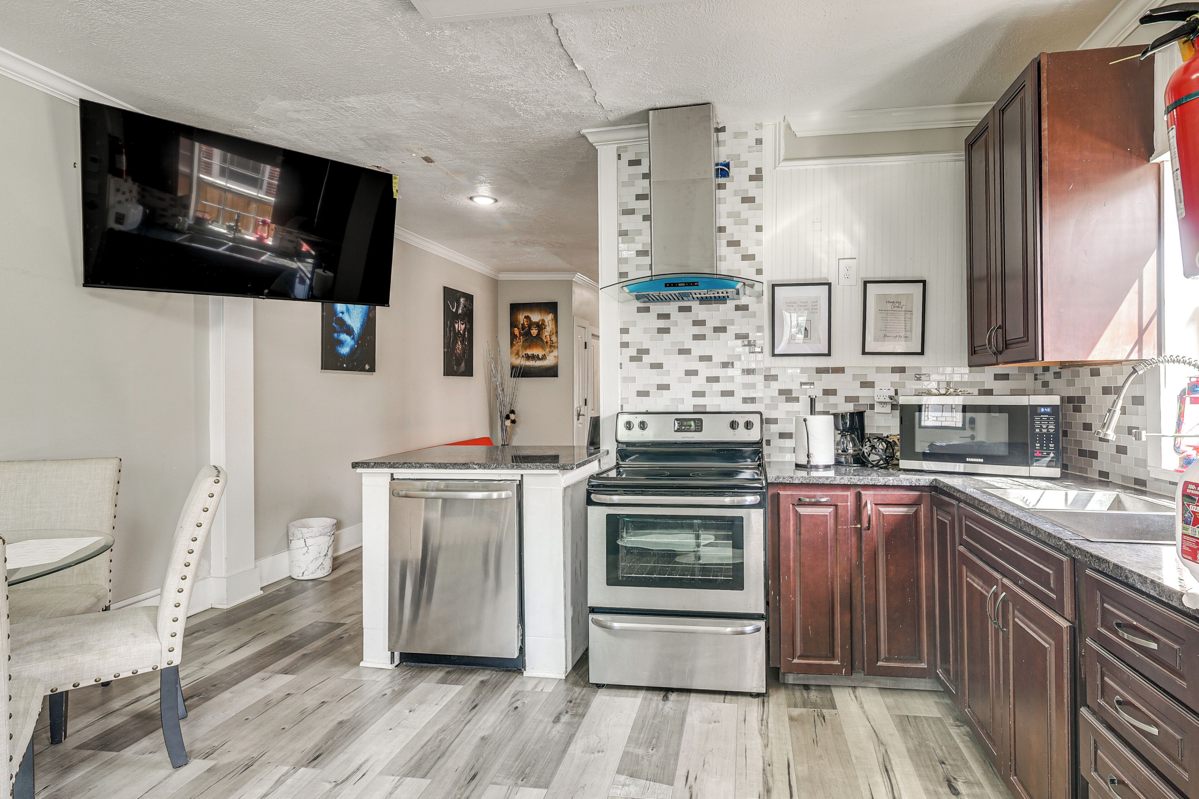 A modern kitchen with stainless steel appliances, a flat-screen TV mounted on the wall, and a tiled backsplash.
